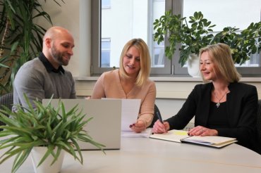 Zwei Frauen und ein Mann blicken in einen Laptop auf einem Bürotisch